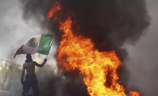 A protester waves a flag as a Waymo taxi burns near the metropolitan detention center of downtown Los Angeles, Sunday, June 8, 2025, following last night's immigration raid protest. (AP Photo/Jae C. Hong)