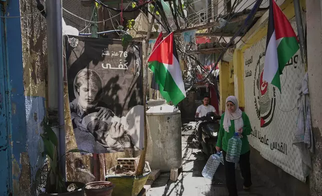A Palestinian woman carries water containers through an alley decorated with Palestinian flags and posters marking the 76th anniversary of the Nakba, in the Mar Elias refugee camp in Beirut, Lebanon, Monday, June 2, 2025. The Arabic writing on the poster reads, "76 years of injustice, enough." (AP Photo/Hassan Ammar)
