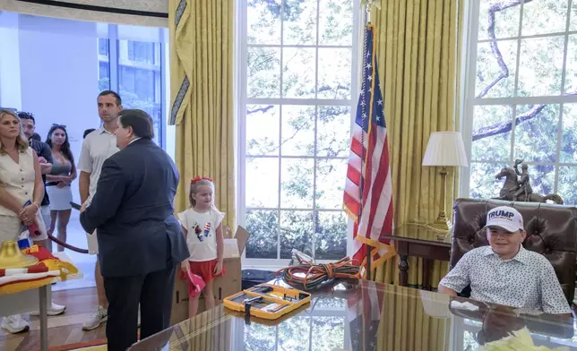 John Michael Cannette,10, right, of Sumrall, Miss., sits behind the Resolute desk as White House Historical Association President Stewart McLaurin, left, talks to a group of tourists, during work to transition a replica of the White House Oval Office from the days of former President Joe Biden with President Donald Trump's decor, at the White House Historical Association in Washington, Wednesday, July 23, 2025. (AP Photo/Rod Lamkey, Jr.)
