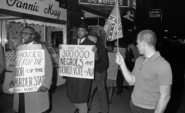 FILE - A man holds a Confederate flag at right, as demonstrators, including one carrying a sign reading, "More than 300,000 Negroes are Denied Vote in Ala.," demonstrate in front of an Indianapolis hotel where Alabama Gov. George Wallace was staying, on April 14, 1964. (AP Photo/Bob Daugherty, File)
