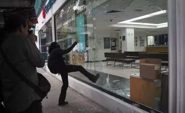 A demonstrator kicks in a bank storefront window at the end of peaceful protest that turned violent against gentrification, as the increase in remote workers has risen prices and increased housing demand in neighborhoods like Condesa and Roma, in Mexico City, Friday, July 4, 2025. (AP Photo/Aurea Del Rosario)