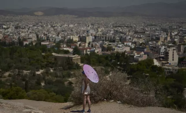 A tourist with an umbrella stands on Filopappou Hill as Athens stretches out in the background, Monday, July 7, 2025, while authorities in Greece impose mandatory work stoppages in parts of the country where temperatures are expected to exceed 40 degrees Celsius (104 Fahrenheit).