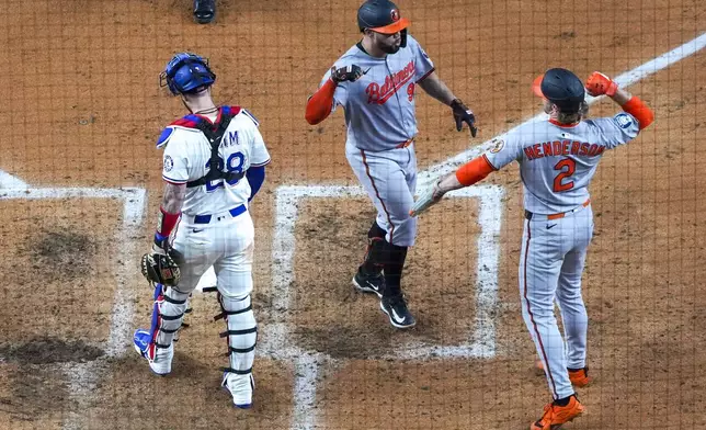 Baltimore Orioles' Gary Sánchez, center, is greeted near home plate by Gunnar Henderson, right, as Texas Rangers catcher Jonah Heim looks on after Sánchez scored both of them on a two-run home run during the fourth inning of a baseball game Tuesday, July 1, 2025, in Arlington, Texas. (AP Photo/Julio Cortez)