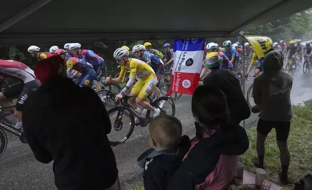 Spectators take cover under an awning as the pack with Slovenia's Tadej Pogacar, wearing the overall leader's yellow jersey, rides in the rain during the twentieth stage of the Tour de France cycling race over 184.2 kilometers (114.5 miles) with start in Nantua and finish in Pontarlier, France, Saturday, July 26, 2025. (AP Photo/Thibault Camus)