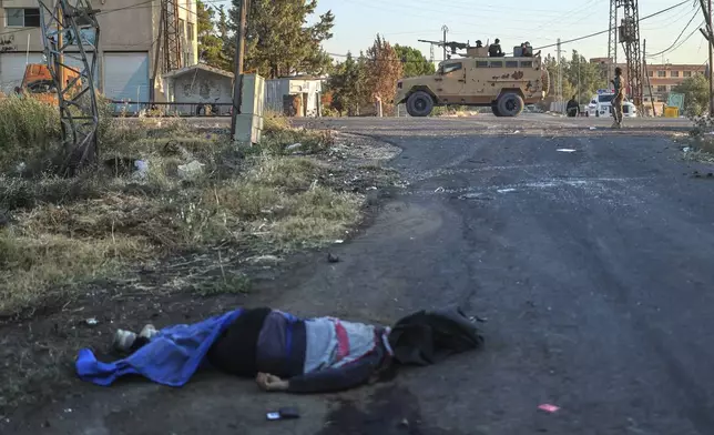 A dead fighter from the Druze militias lies on the ground, as Syrian government forces, background, deploy at Mazraa village on the outskirts of the city of Sweida, where clashes erupted between Sunni Bedouin clans and Druze militias, southern Syria, Monday, July 14, 2025. (AP Photo/Ghaith Alsayed)