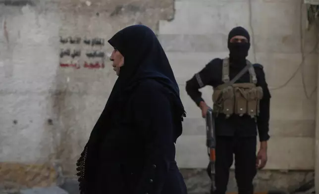 A Syrian woman walk past a Syrian government security man, outside the shrine of al-Sayydah Zeinab, the grand daughter of Prophet Mohammed, where Shiite worshippers attend Ashoura ritual, south of Damascus, Syria, Thursday, July 3, 2025. (AP Photo/Omar Sanadiki)