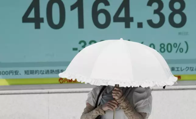 A person walks under the intense sun near an electronic stock board displaying Japan's Nikkei index at a securities firm Tuesday, July 1, 2025, in Tokyo. (AP Photo/Eugene Hoshiko)