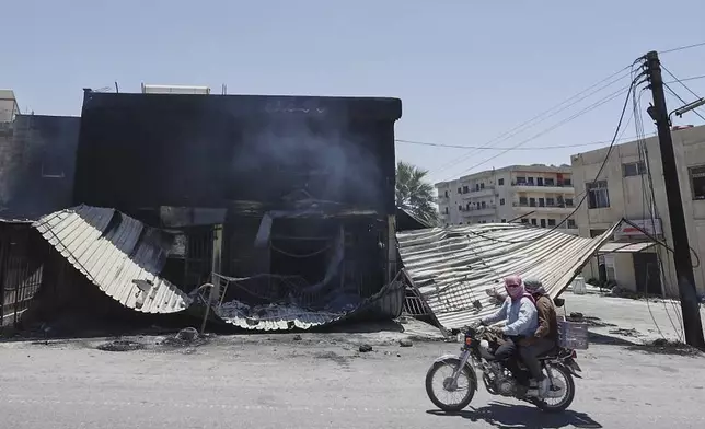 Syrian citizens ride in front of a shop that was burned during clashes between Syrian government forces and Druze militias on the outskirts of Sweida city, southern Syria, Wednesday, July 16, 2025. (AP Photo/Omar Sanadiki)