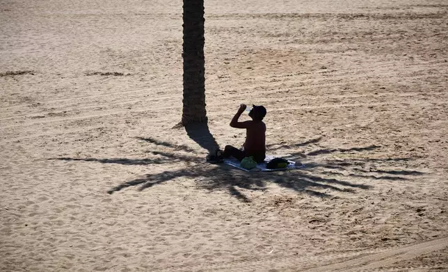 A man takes advantage of the shade of a palm tree to protect himself from the sun while hydrating himself by drinking water, on a hot day at the beach in Barcelona, Spain, Wednesday, July 2, 2025. (AP Photo/Emilio Morenatti)
