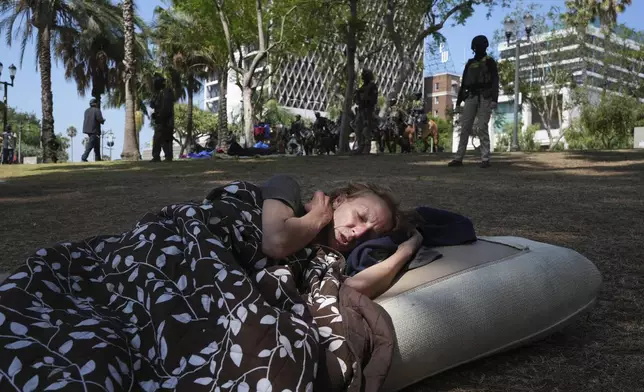 A person sleeps on the ground in front of federal agents stage at MacArthur Park Monday, July 7, 2025, in Los Angeles. (AP Photo/Damian Dovarganes)