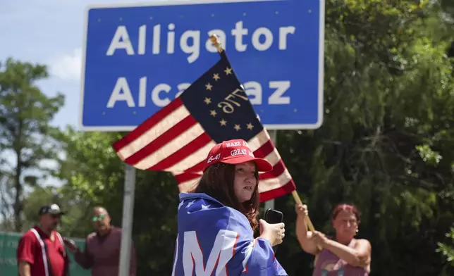 Dianne Mourer waves an American flag as Rana Mourer stands in front of a sign reading "Alligator Alcatraz" outside the Dade-Collier Training and Transition Facility, Saturday, July 12, 2025, in Ochopee, Fla. (AP Photo/Alexandra Rodriguez)