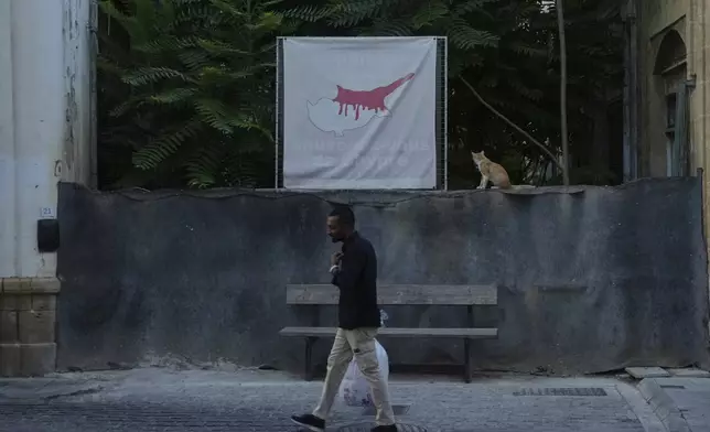 A man walks in front of a blocked road across the U.N buffer zone as a cat sits next of a banner shows the Cyprus island divided, the Turkish occupied area at the north and Cyprus republic at the south, in divided capital Nicosia, Cyprus, Thursday, July 17, 2025. (AP Photo/Petros Karadjias)