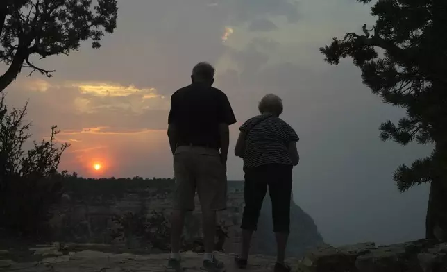The setting sun peeks through the smoke from the Dragon Bravo Fire as it moves over the Grand Canyon from the North Rim as two tourists take in the scene from a South Rim overlook Monday, July 14, 2025, in Grand Canyon National Park, Ariz. (AP Photo/Ross D. Franklin)