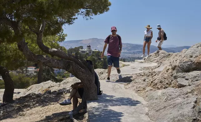 A woman sits in the shade of a tree as other tourists walk on Filopappou Hill in Athens, Tuesday, July 8, 2025. (AP Photo/Petros Giannakouris)