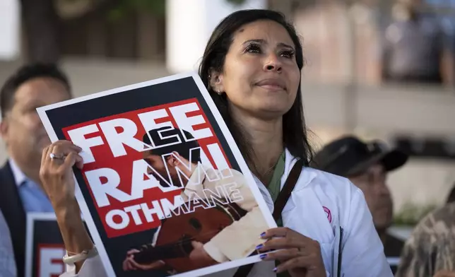 Dr. Wafaa Alrashid, center, whose husband, Rami Othmane, a Tunisian musician, is detained at the U.S. Immigration and Customs Enforcement facility, holds a sign during a rally outside the facility in Los Angeles Friday, July 25, 2025. (AP Photo/Jae C. Hong)