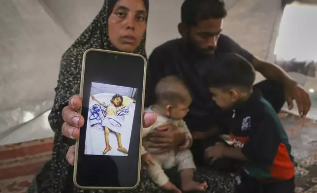 Sitting next to her husband, Amin, and two of their children, Fatma Mashi shows a photo on her cellphone of the body of her 2½-year-old daughter, Ro'a, who doctors say had no preexisting conditions and died of severe malnourishment, at their tent in Muwasi, southern Gaza Strip, Friday, Aug. 8, 2025. Experts say it's not unusual for one family member to fare worse than the others. (AP Photo/Mariam Dagga)