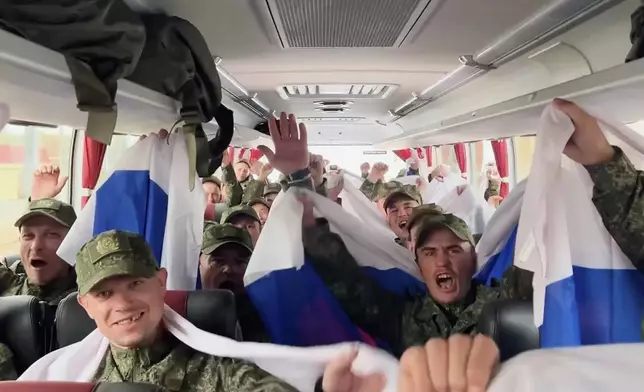 In this photo, taken from a video and distributed by the Russian Defense Ministry Press Service on Sunday, Aug. 24, 2025, Russian servicemen wave Russian national flags sitting in a bus at an exchange area in Belarus after returning from captivity during a POWs exchange of a group of servicemen between Russia and Ukraine. (Russian Defense Ministry Press Service via AP)