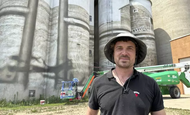 Artist Guido van Helten poses for a photo on Monday, July 14, 2025, in front of the grain elevator and silos in Minot, N.D., where he is painting a large mural. Behind him is the boom lift he uses to ascend the structure. (AP Photo/Jack Dura)