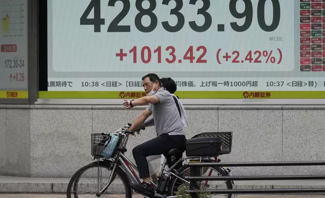 Two persons ride bicycles in front of an electronic stock board showing Japan's Nikkei index at a securities firm Tuesday, Aug. 12, 2025, in Tokyo. (AP Photo/Eugene Hoshiko)