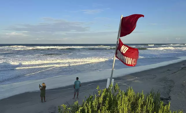 As waves from Hurricane Erin crash and “No Swimming” flags flap in the breeze, people walk on the beach in Nags Head, N.C., on Tuesday, Aug. 19, 2025. (AP Photo/Allen G. Breed)