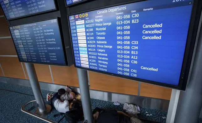 People sleep under a flight information board at the Vancouver International Airport in Richmond, B.C., Canada, on Saturday, Aug. 16, 2025. (Ethan Cairns/The Canadian Press via AP)
