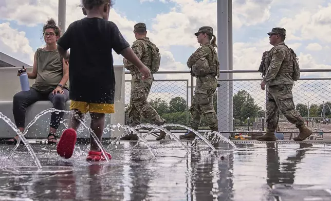 Armed members of the Ohio National Guard pass by children playing in a water feature at The Wharf, Monday, Aug. 25, 2025, in Washington. (AP Photo/Jacquelyn Martin)
