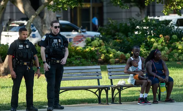 A family sits on a bench in Franklin Park in downtown Washington as Washington Metropolitan Police patrol Thursday, Aug. 14, 2025. (AP Photo/Cliff Owen)