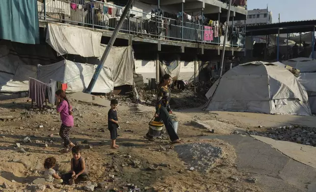 A Palestinian woman carries buckets as she walks past tents for displaced people in a school used as a shelter in Gaza City, Wednesday, Aug. 27, 2025. (AP Photo/Jehad Alshrafi)