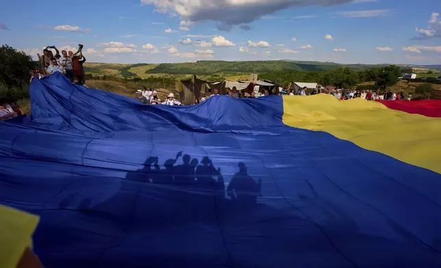 People cast shadows on a Romanian flag they unfurled in Poiana, Romania, Saturday, July 12, 2025, during the Getodava historical reenactment festival. (AP Photo/Andreea Alexandru)
