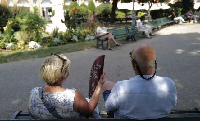 People sit on a bench in a park and use a fan Tuesday, Aug. 12, 2025 in Toulouse, southwestern France. (AP Photo/Fred Scheiber)