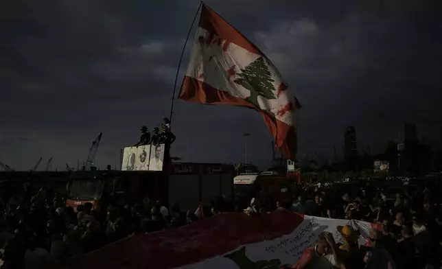 Relatives of victims of the deadly 2020 Beirut port explosion stand on the ladder of a firetruck, holding portraits of their loved ones and a giant Lebanese flag, during a gathering marking the fifth anniversary of the massive blast, outside the port of Beirut, Lebanon, Monday, Aug. 4, 2025. (AP Photo/Hassan Ammar)