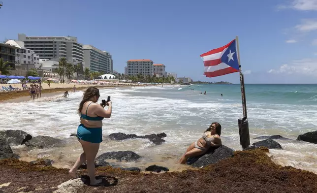 A woman poses for photos next to a Puerto Rican flag along the beach in Condado, Puerto Rico, as Hurricane Erin approaches, Friday, Aug. 15, 2025. (AP Photo/Alejandro Granadillo)