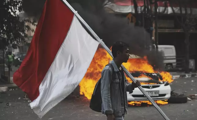 A man carries an Indonesian national flag near a burning car during a protest following the death of a delivery rider in clashes between riot police and students protesting against lawmakers' allowances, in Jakarta, Indonesia, Friday, Aug. 29, 2025. (AP Photo/Achmad Ibrahim)