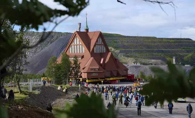 People gather outside the Kiruna Church, a Sami style wooden Swedish Lutheran church, called Kiruna Kyrka in Swedish, in Kiruna, Sweden, Tuesday, Aug. 19, 2025, during its move along a 5-kilometer (3-mile) route east to a new city center as part of the town's relocation. (AP Photo/Malin Haarala)
