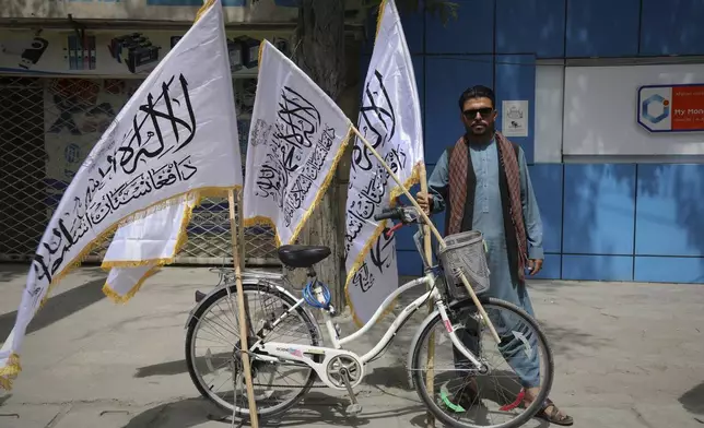 A man poses for the camera next to his bicycle while selling Taliban flags ahead of celebrations marking the fourth anniversary of the U.S. withdrawal from Afghanistan and the beginning of Taliban rule in Kabul, Afghanistan, Thursday, Aug. 14, 2025. (AP Photo/Siddiqullah Alizai)