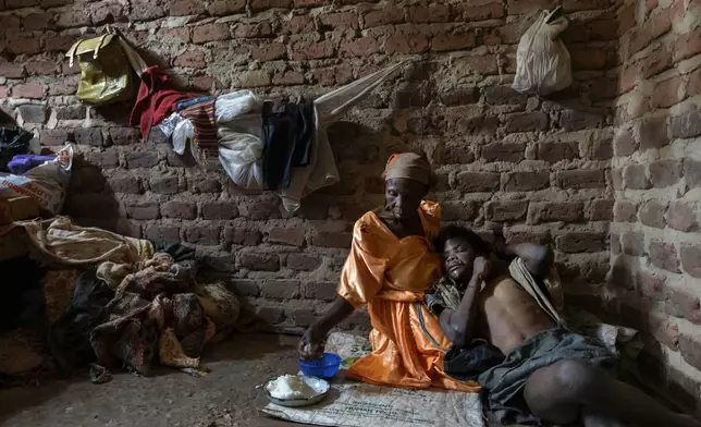 Alice Mary Nasanga, who is about 70 and was banished by members of her family after she was accused of witchery, holds her disabled son, Ibrahim Kamya, 30, as she feeds him on the floor of the home they share in Nabalanga, Uganda, Saturday, Nov. 16, 2024. "There is no one. Only God knows," she says of what will happen to him when she's physically no longer able to care for him. (AP Photo/David Goldman)