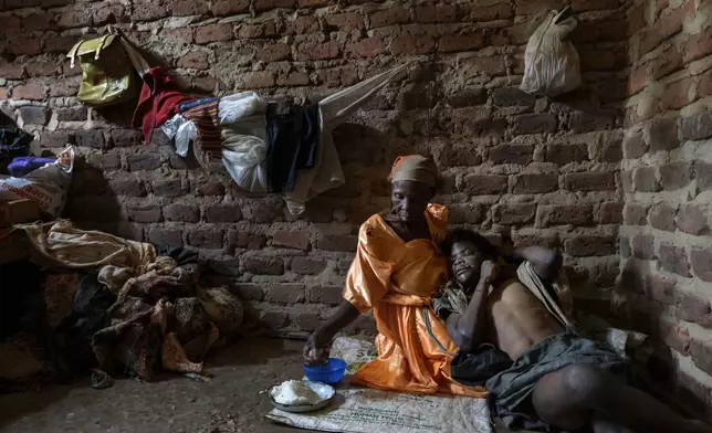 Alice Mary Nasanga, who is about 70 and was banished by members of her family after she was accused of witchery, holds her disabled son, Ibrahim Kamya, 30, as she feeds him on the floor of the home they share in Nabalanga, Uganda, Saturday, Nov. 16, 2024. (AP Photo/David Goldman)