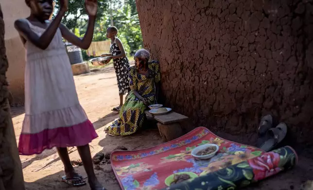 Rose Liru, 94, right, eats dinner prepared by her granddaughter, Parvin Nakawesi, 9, left, and grandniece, Brenda Mungulu, 11, rear, at the home they share, Friday, Nov. 22, 2024, in Magogo, Uganda. (AP Photo/David Goldman)