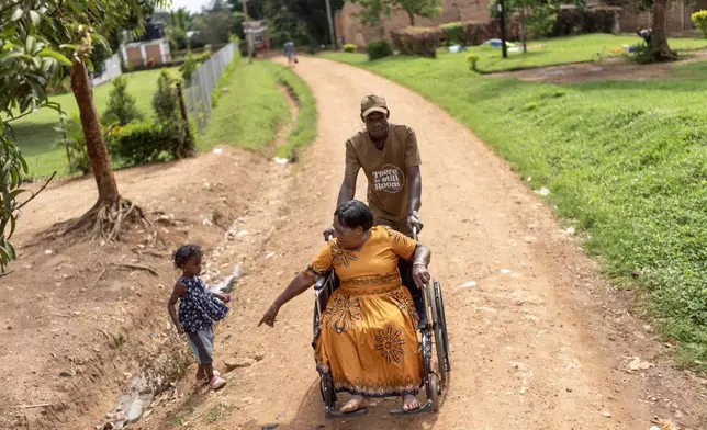 Norah Kasozi, 73, gestures to her granddaughter, Shalom, 4, to be careful near a ditch as she's pushed in a wheelchair to her home by Isaac Okware following a physical therapy visit to a nearby clinic, Saturday, Nov. 16, 2024, in Mukono, Uganda. (AP Photo/David Goldman)