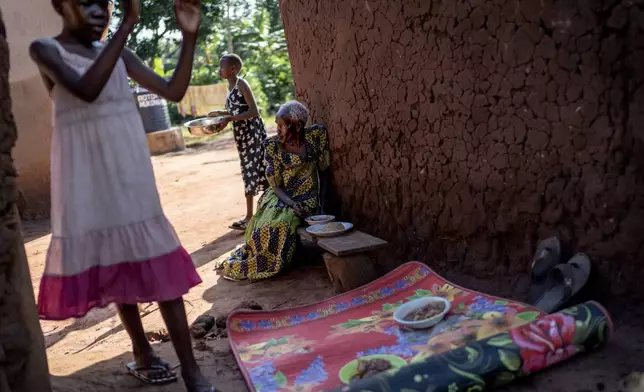 Rose Liru, 94, right, eats dinner prepared by her granddaughter, Parvin Nakawesi, 9, left, and grandniece, Brenda Mungulu, 11, rear, at the home they share, Friday, Nov. 22, 2024, in Magogo, Uganda. (AP Photo/David Goldman)