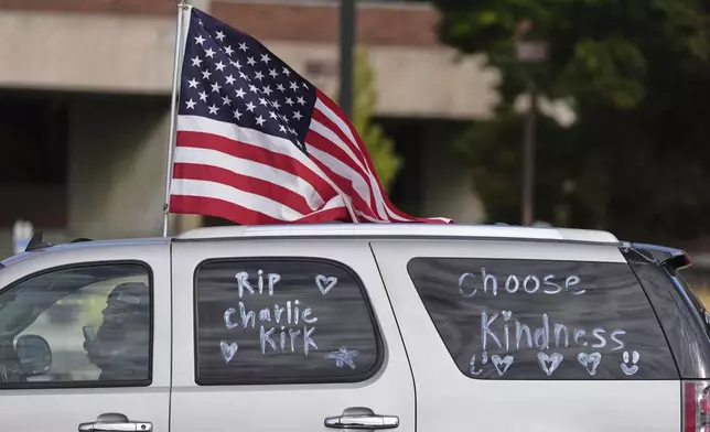 A vehicle marked with messages written on its windows in tribute to Turning Point USA CEO and co-founder Charlie Kirk and carrying an American flag, drive past at Utah Valley University, Saturday, Sept. 13, 2025, in Orem, Utah. (AP Photo/Lindsey Wasson)