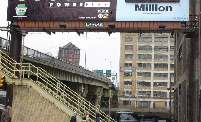 FILE - People move along the sidewalk at lunchtime in Pittsburgh in front of a billboard advertising the current Powerball Jackpot on Monday, March 21, 2005. (AP Photo/Keith Srakocic, File)