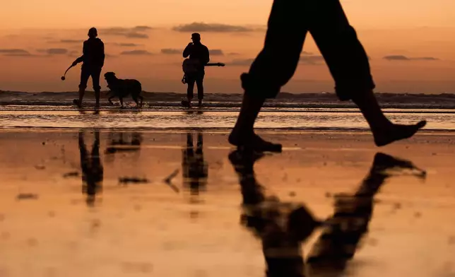 Chris Strong, center, plays a guitar as his spouse Mary Strong, left, throws a tennis ball for their dog as the sun sets behind Ocean Beach's dog beach Tuesday, Oct. 7, 2025, in San Diego. (AP Photo/Gregory Bull)