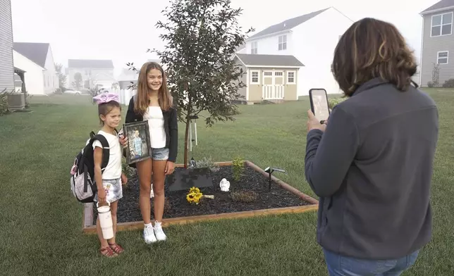 Ashlee Dahlberg takes photos of her daughters Ava, left, and Khloe, on the first day of school in front a memorial for their brother, Liam, in their yard in Lowell, Ind., on Wednesday, Aug. 13, 2025. (AP Photo/Laura Bargfeld)