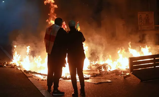 Demonstrators with a Georgian and Ukrainian national flags stand behind a burning barricade not far from a police line during an opposition rally in the city center of Tbilisi, Georgia, on Saturday, Oct. 4, 2025, boycotting the municipal elections and calling for the release of political opponents. (AP Photo/Zurab Tsertsvadze)