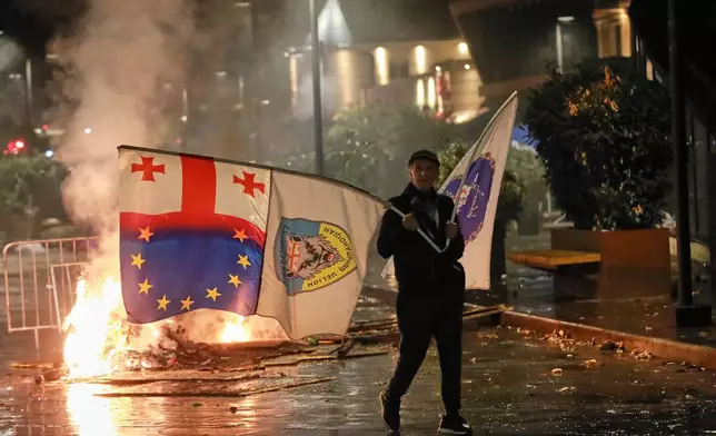 A demonstrator with a Georgian national, EU and other flags walks in front of police line during an opposition rally in the city center of Tbilisi, Georgia, on Saturday, Oct. 4, 2025, boycotting the municipal elections and calling for the release of political opponents. (AP Photo/Zurab Tsertsvadze)