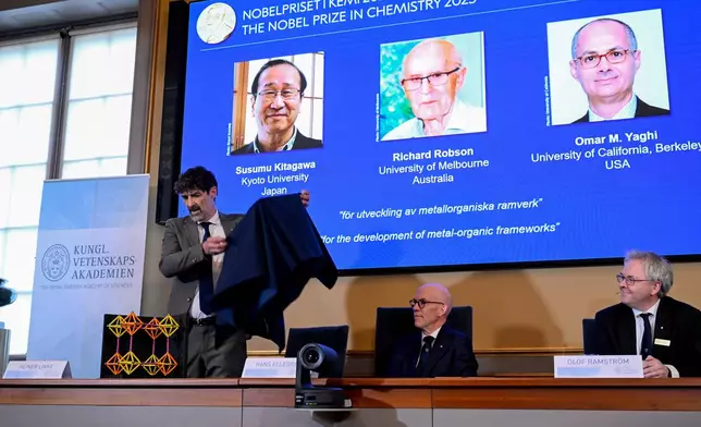 Chair of the Nobel Committee for Chemistry Heiner Linke makes a demonstration, next to Secretary General of the Swedish Academy of Sciences Hans Ellegren, and Member of the Nobel Committee for Chemistry Olof Ramstrom, right, after they announce Susumu Kitagawa, Richard Robson and Omar Yaghi, on screen behind, as the recipients the Nobel Prize in Chemistry, at the Nobel Assembly of the Karolinska Institutet, in Stockholm, Sweden, Wednesday, Oct. 8, 2025. (Fredrik Sandberg/TT News Agency via AP)
