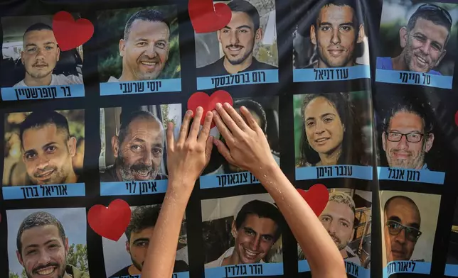 Hands paste a heart-shaped sticker onto a banner with pictures of Israeli hostages during a a gathering at a plaza known as hostages square in Tel Aviv, Israel, Monday, Oct. 13, 2025. (AP Photo/Oded Balilty)