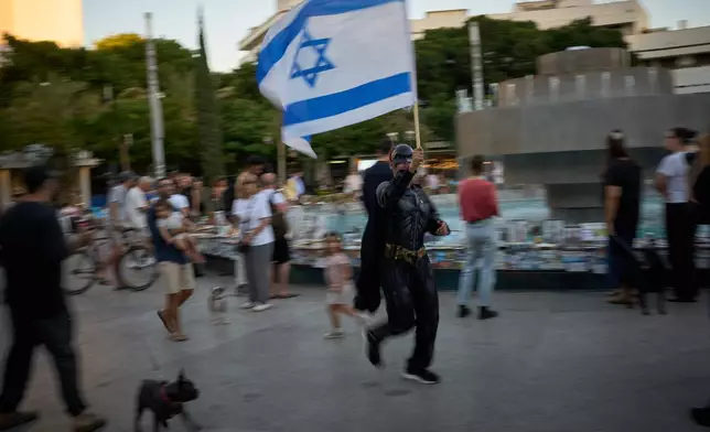 A man dressed as Batman waves an Israeli flag as people watch photos of Israelis who were killed in the Oct. 7, 2023 deadly Hamas attack on Israel, at a memorial marking two years anniversary of the assault, in Tel Aviv, Israel, Tuesday, Oct. 7, 2025. (AP Photo/Emilio Morenatti)
