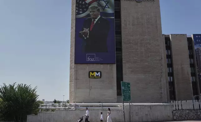People walk past large outdoor screen displaying an image depicting the U.S. President Donald Trump, in Jerusalem, Saturday, Oct. 11, 2025, after Israel and Hamas agreed to a pause in their war and the release of the remaining hostages. (AP Photo/Mahmoud Illean)